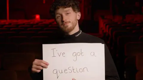 Jacob Molloy/Woolton Cinema A screengrab from the big screen showing Jacob holding a placard that says "I've got a question". Jacob has brown curly hair and similar coloured moustache and a stubbly beard. 