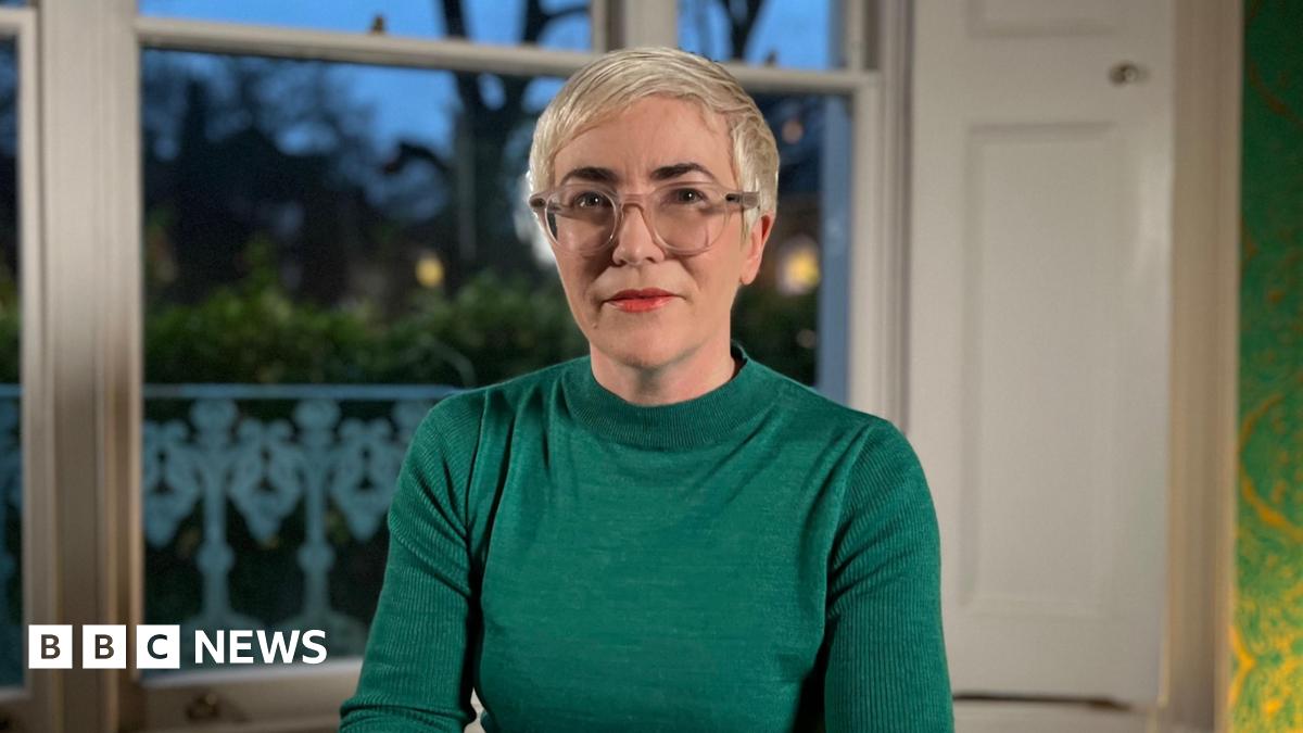 Alice Sullivan sits in a room looking at the camera. She has short platinum blonde hair, glasses and bright lipstick. She's wearing an emerald green jumper.