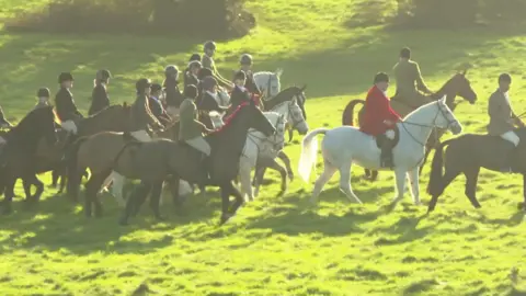 BBC A group of people riding horses through a grassy field. 