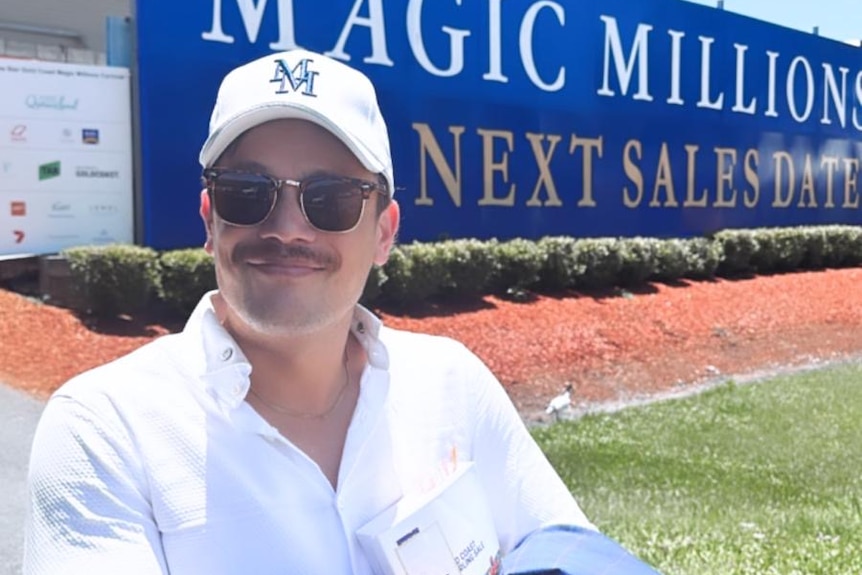 A man in sunnies and a cap standing in front of a Magic Millions sign.
