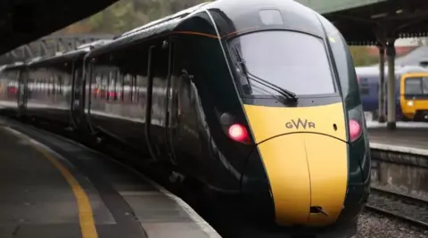 PA Media A GWR train pulling into a station. The train is dark green with a white front at the GWR logo on the front. An older train can be seen at another platform in the background