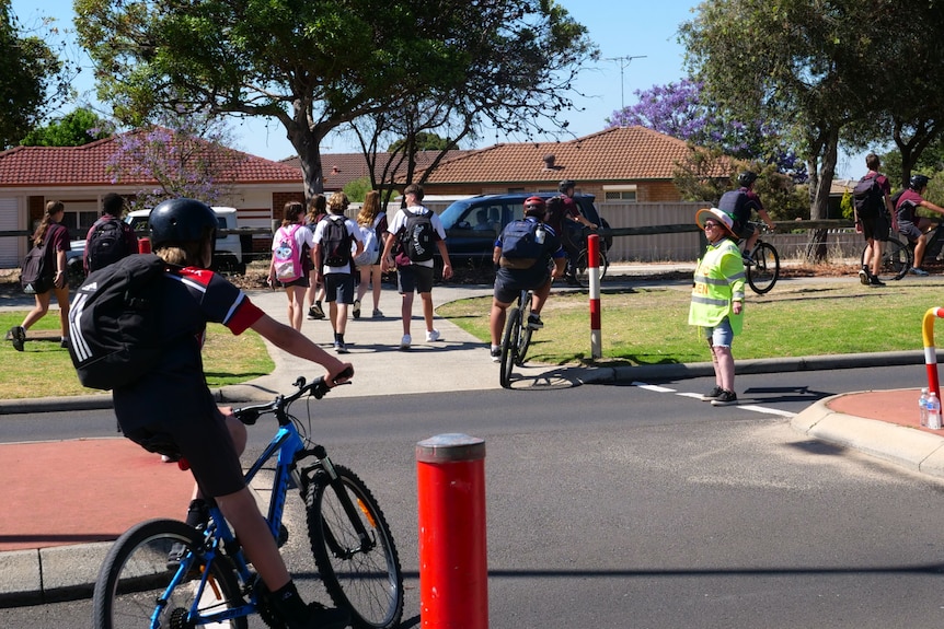 School students walk and ride bikes across a road while a traffic warden watches on.
