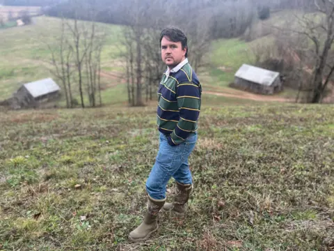 BBC/Ellie House A man on a hill looks at the camera, wearing jeans and boots, there are barns and rolling hills in the distance.