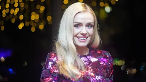 PA News Katherine Jenkins wearing pink sequinned top and smiling at the camera. She has long blonde hair worn down and the backdrop to the photo is dark, with some twinkly lighting