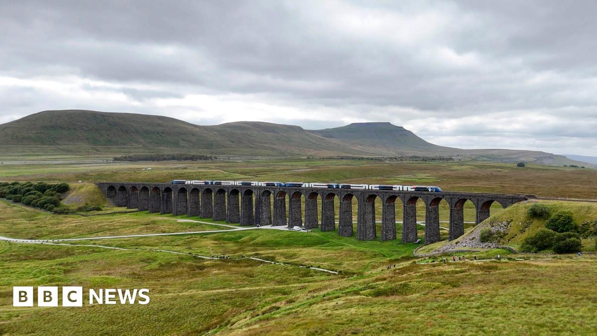A train running along the Settle to Carlisle railway line over the Ribblehead Viaduct. The stone viaduct with its many arches is surrounded by rolling green land with fells in the background.