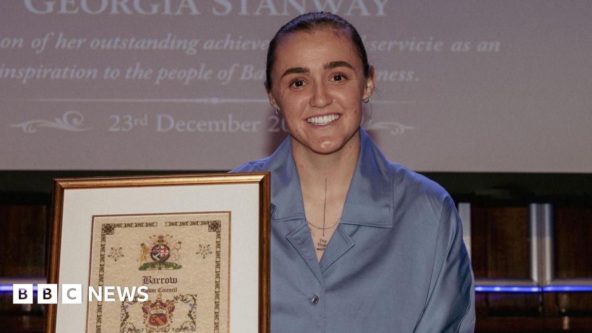 Georgia Stanway, who has brown hair, in a blue blouse wearing a necklace. She is smiling and holding a certificate declaring her 'Freedom of the Town' award.