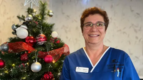 Helena is dressed in a blue uniform with a name badge and pens in the pocket stands beside a Christmas tree decorated with red ribbons, baubles, and a star topper. The setting includes a light-colored wall with framed artwork.
