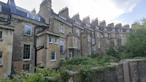 Google A row of three-storey brick terraced houses in Bath. They are mostly built using the Cotswold stone style of the area and there are plants and bushes running along a wall in front of the properties.