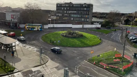 Bedford Borough Council Shot from above of a roundabout covered in grass, with a sculpture in the centre. There are cars going round the roundabout. There is a six-storey building on the far side of the roundabout, and smaller buildings to the left.