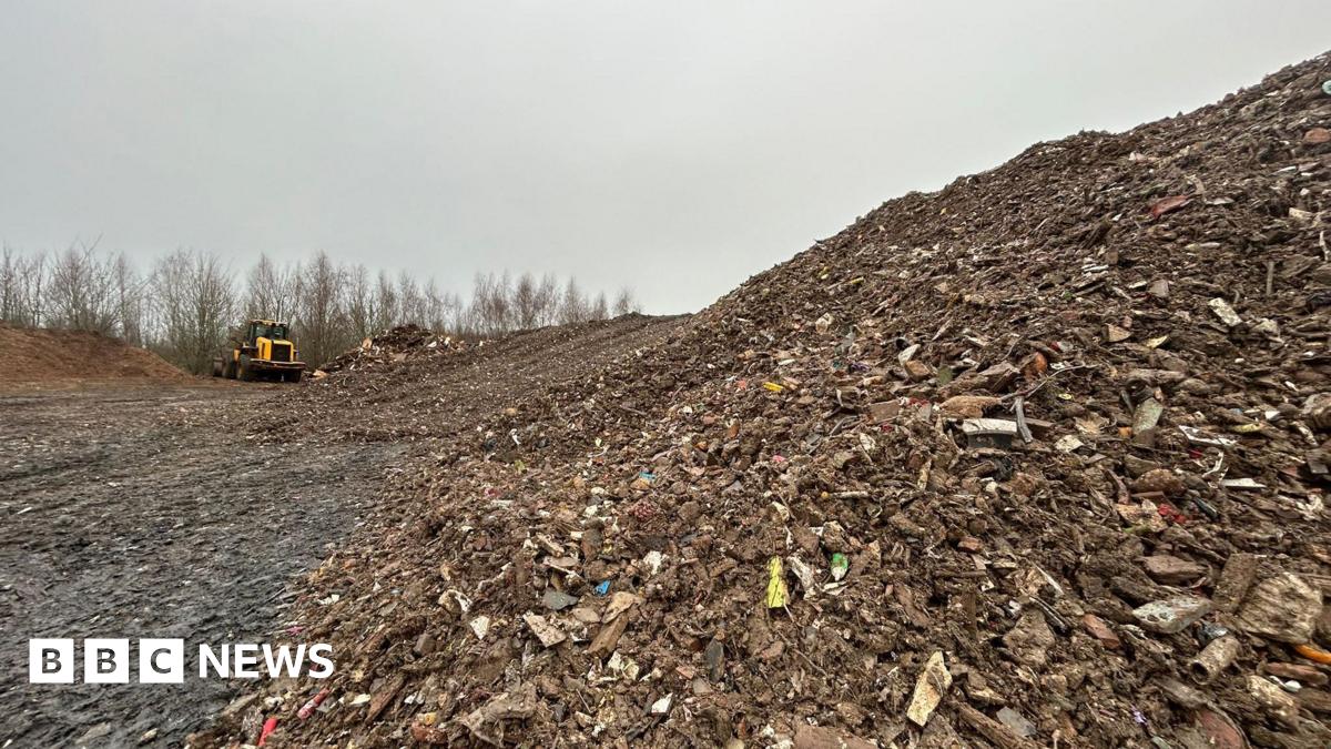 A close up image of a large pile of waste which extends out into the distance of the photograph. The waste is made up of wood, plastic and other bits of household waste. There is a yellow JCB truck in the background.