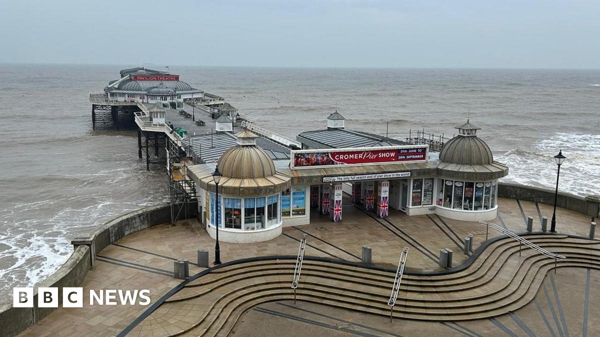 Cromer pier viewed from land and an elevated position directly in front of it. The jetty has white railings on both sides, benches in the centre and a grey-roofed building at the far end. The entrance is flanked by circular, glass fronted buildings with pointed, dome roofs.  The sea is a grey-brown, the sky a blue-grey.