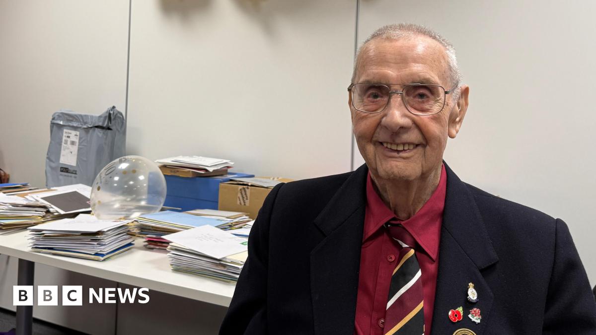 An elderly man with short white hair, large wire-framed glasses and a fairly narrow face smiles at the camera. He wears a red shirt, red, black, white and yellow tie, and black blazer with various poppy and remembrance badges. Behind him on a table are many stacks of cards in envelopes.