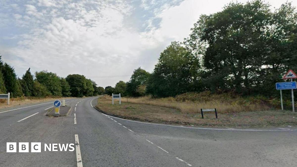 A Google street view image of a road junction. A single carriageway can bee seen with traffic island in the centre and two white signposts either side on grassy verges, lined by trees. A turning to the right is signed Rocky Lane and a sign warns of people riding horses in the area.