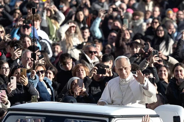 A religious leader waves to a crowd from a vehicle, surrounded by people taking photos during an outdoor event