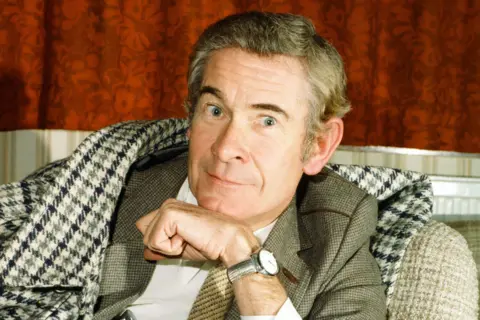 Getty Images Stanley Baxter is seated indoors wearing a checked overcoat draped over the shoulders and a light brown suit jacket underneath. He has a light-coloured dress shirt and a wristwatch with a round face on the left wrist. One hand is resting near the chin in a thoughtful pose. The background features patterned orange curtains and part of a white radiator.
