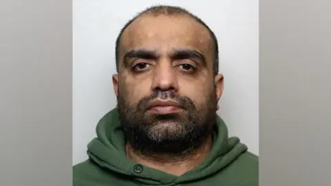 West Yorkshire Police A police mugshot of Sharaz Ali. He has short, fuzzy black hair and a a beard. He is wearing a green hooded top.