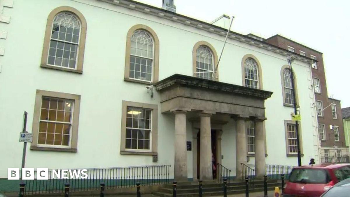 Enniskillen Courthouse. The building is white with stone window surrounds. It has four concrete pillars at the front door. There are black bollards outside. A red car is driving past. Behind the court is a brick building.