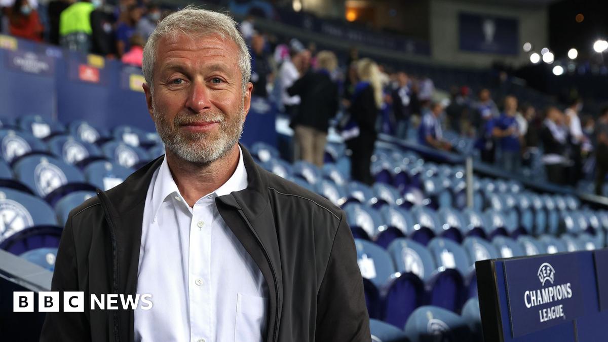 Roman Abramovic, a man in his late 50s, standing in front of football stadium seating. He is wearing a white shirt and a black casual jacket