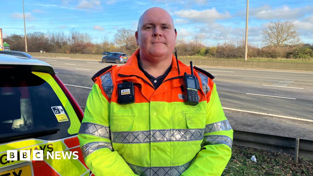 Traffic officer Robert Oates is standing by the M1. There are cars behind him on the motorway. He is wearing a fluorescent jacket with a radio and a bodycam attached to it.