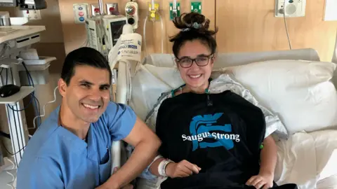 Mia Tretta Mia Tretta is in a hospital bed wearing hospital gown with a black top draped over it that says Saugus Strong in white letters. A surgeon sits next to her in blue surgical scrubs. Both of them are smiling. 