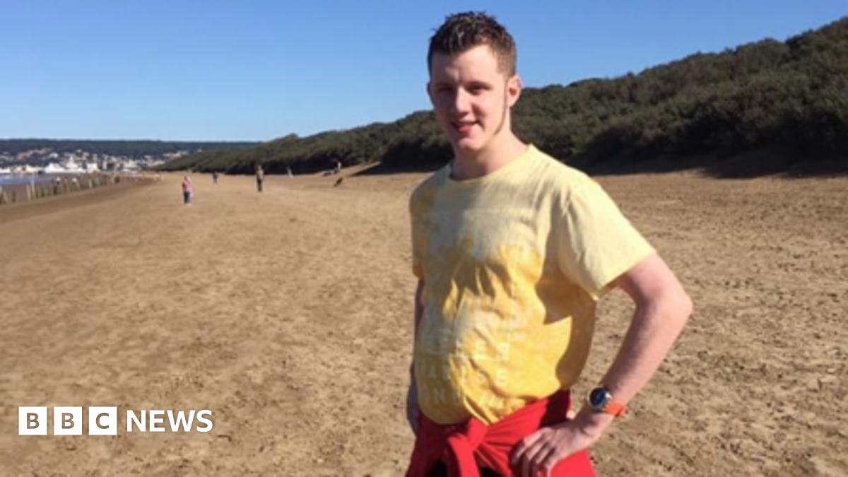 Jamie Cole is pictured standing on a beach wearing a yellow T-shirt. He has a red sweater tied around his waist and he is standing with his hands on his hips. He has short dark blonde hair. People can be seen in the background.