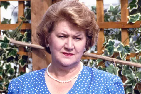 Patricia Routledge is wearing a blue dress with a white polka dot pattern and a single-strand pearl necklace. The background features a wooden trellis adorned with green ivy leaves, suggesting an outdoor garden or patio setting. The lighting appears natural, indicating daytime.