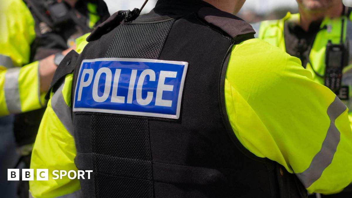 Close-up of 'POLICE' marking written on the back of a hi-visibility stab proof vest worn by a trio of police officers at the scene of an incident.