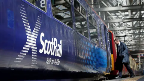 PA Media A navy blue ScotRail train at an indoor train station during the day as a passenger in business dress boards the train with a three-carriage red and cream train in the background