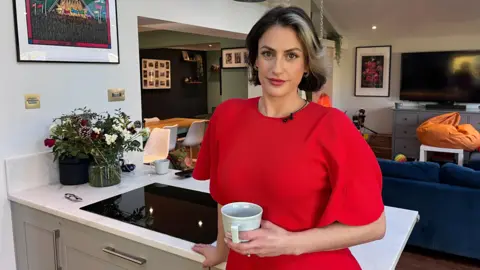 Madelaine Thomas is standing in her kitchen holding a mug and smiling at the camera. She is wearing a red short-sleeved dress. She has short dark hair with a blonde streak at the front. Behind her is a navy sofa, a dining table and walls covered in framed photos and artwork. Her hand rests on a kitchen unit which has a built in hob. There are also flowers in vases on the unit - to the left of the image.