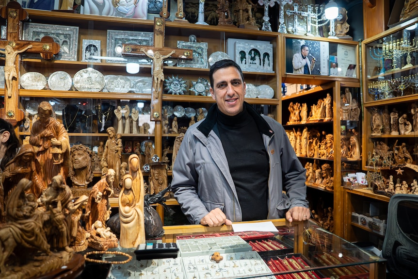 A man stands behind a counter with christian figurines on the side and in shelves behind him. 