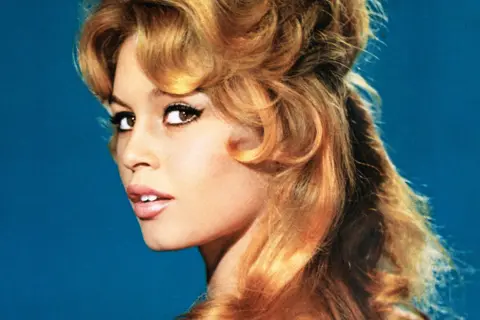Getty Images A young Brigitte Bardot has her head turned as she looks at the camera. She has long blond hair in loose curls, light pink lipstick and dark eye mascara. She is set against a blue background.