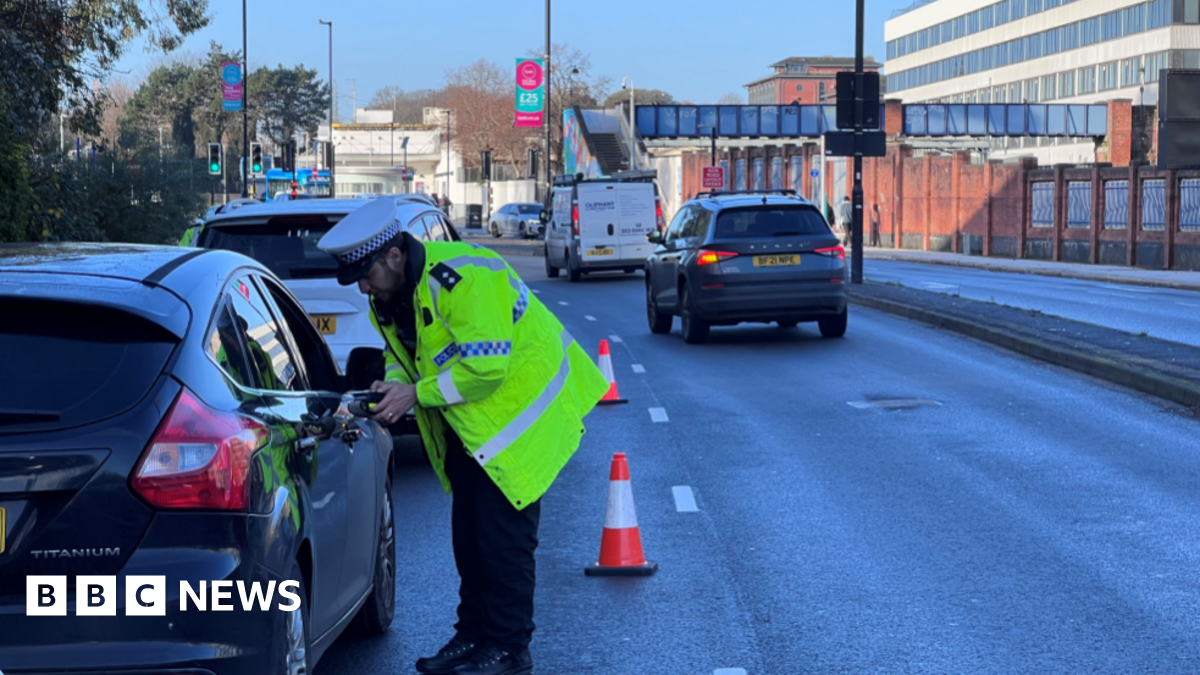 A male police officer in a hi-viz yellow jacket wearing white hat has stopped by a black car to check if they are driving impaired.