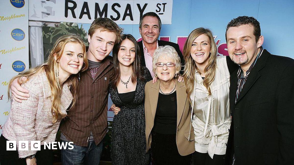 Seven people stand with their arms around each other's shoulders in front of a partition with the graphic of a street sign saying "Ramsay St"
