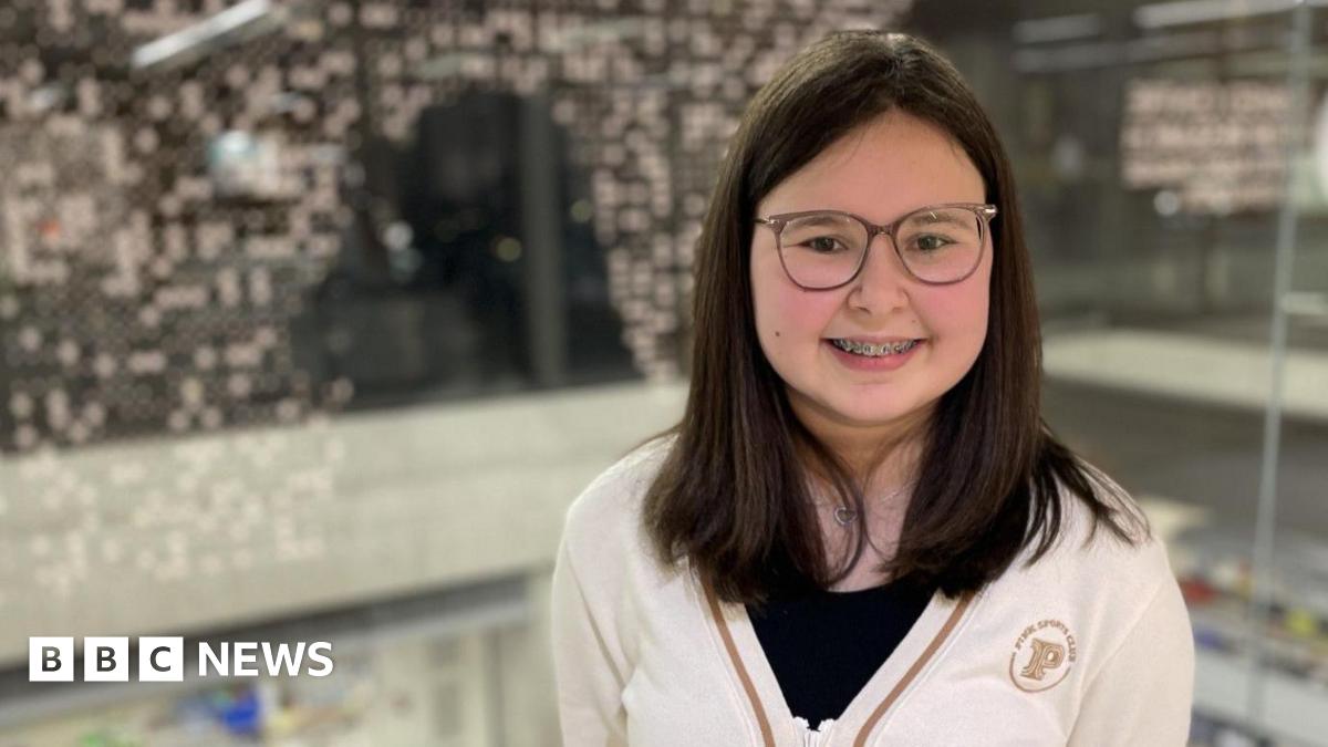 A teenage girl with dark hair smiles at the camera, wearing a white cardigan