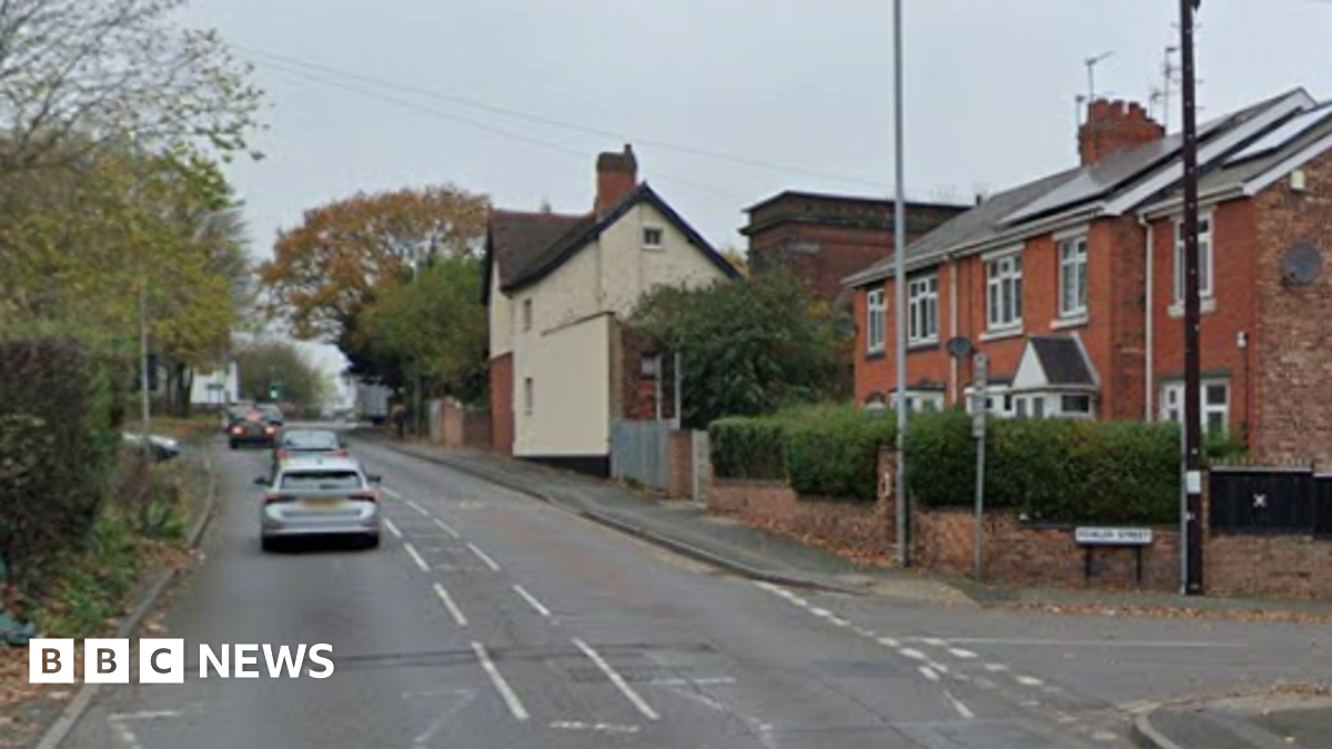 Cars drive up the main road on the left and houses can be seen on the right side of the road before a turning for Fowler Street.