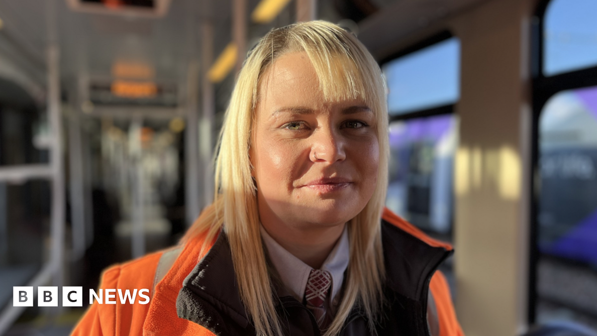 Head and shoulders image of Holly Thompson on board a tram. She has long blonde hair and is wearing a high-vis orange jacket over a shirt and tie. She has a silver nose-ring.