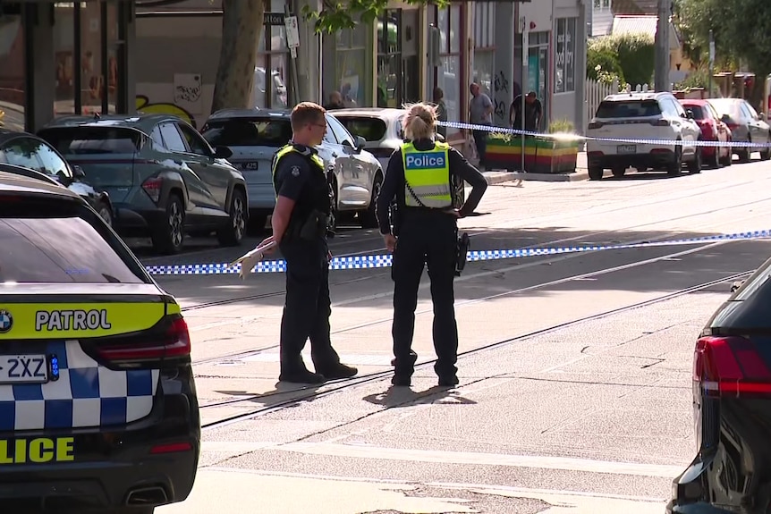 Police officers stand behind police tape next to a police car.