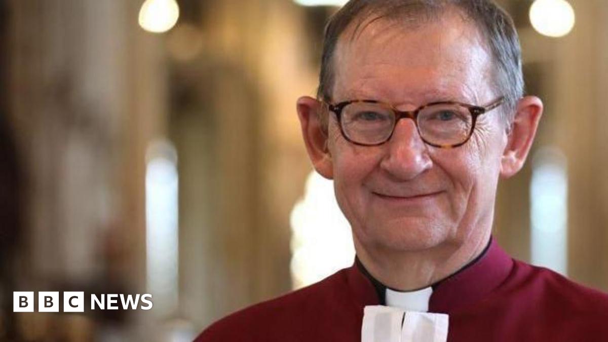 A smiling Chris Dalliston looks directly at the camera. He is wearing tortoise shell spectacles and has short grey hair. He is wearing a red shirt with a white clerical collar.  He is standing in a cathedral.