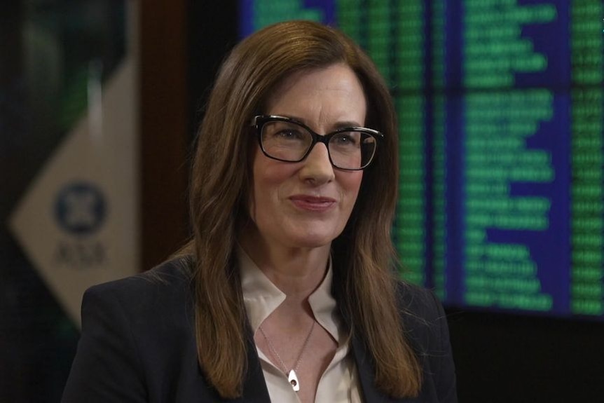 A middle-aged woman with glasses and long hair listens in a TV interview at the stock exchange