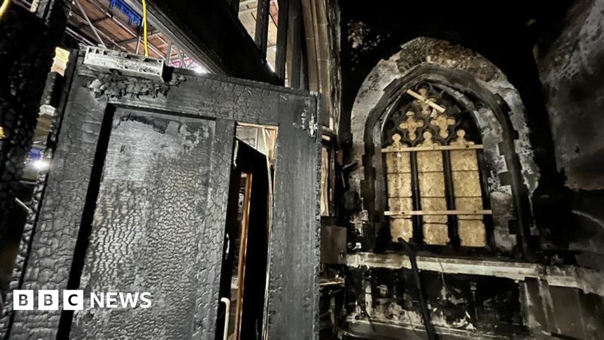 Damage to the inside of a church's kitchen where a fire was accidentally started. The room is coated in thick soot and there is fire and heat damage to the walls and door of the room, including a boarded up stained glass window.