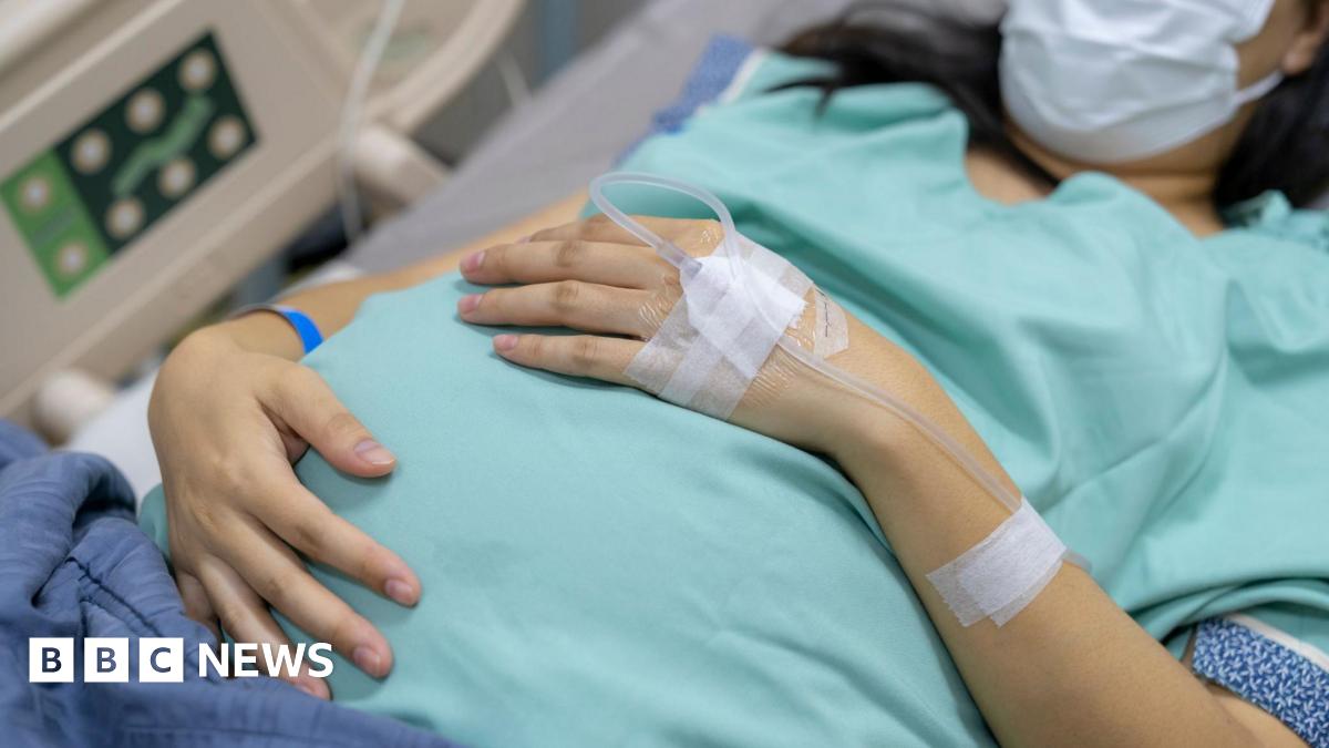 A pregnant woman waits to give birth lying in a hospital bed, wearing a blue gown and mask, with a tube attached to the back of her hand
