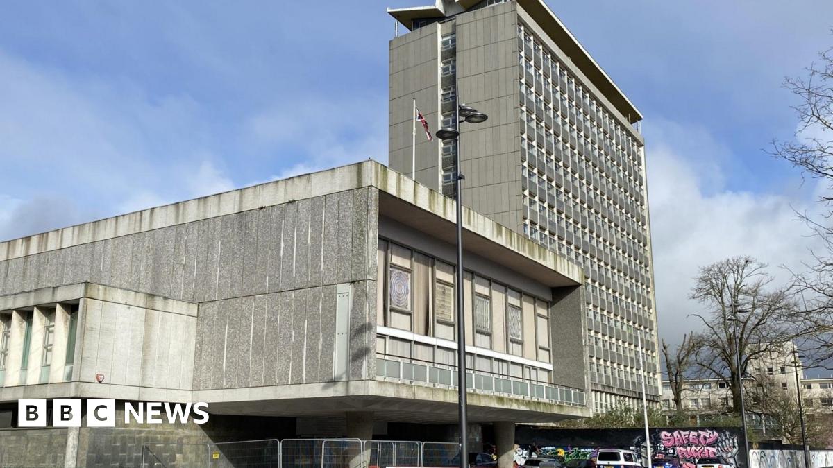 A grey tower block with a lower grey 1960s building to its left is photographed against a pale blue sky. A Union Flag is flying form a flagpole on the lower building.
