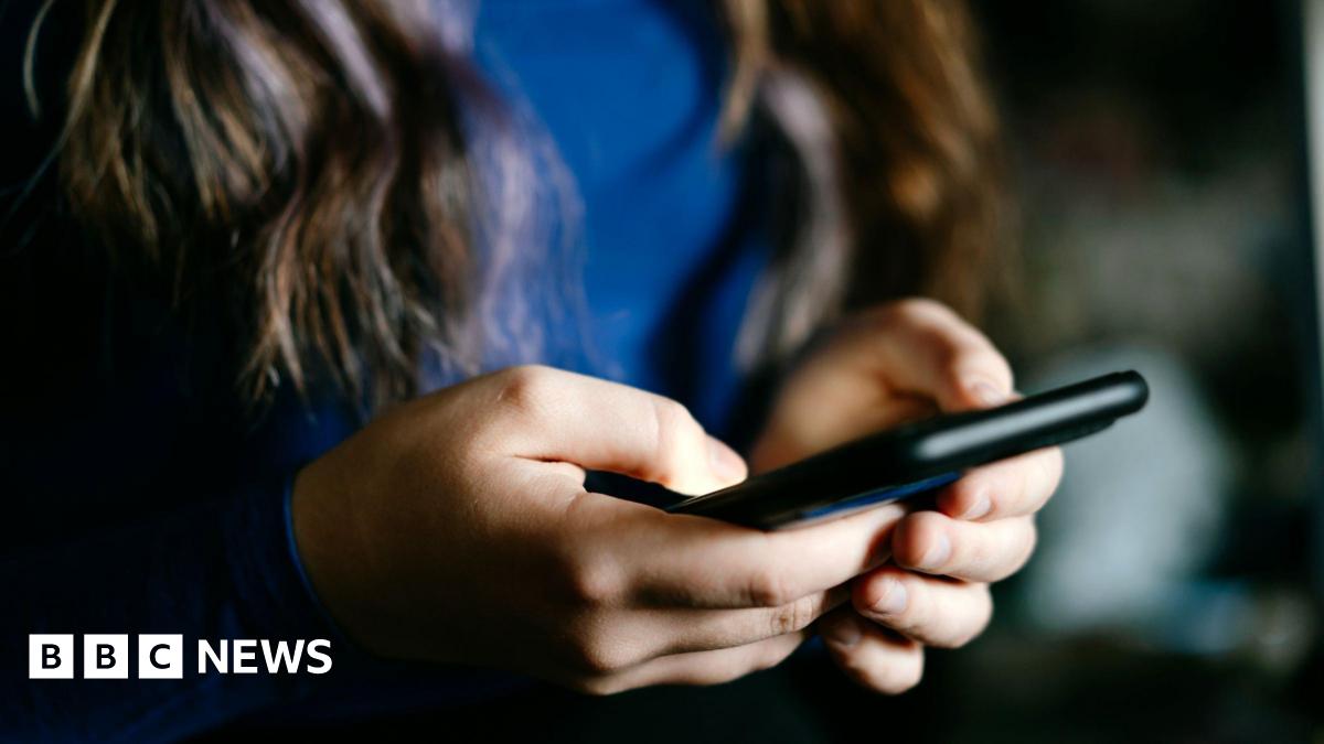 A girl with long brown hair and wearing a blue top holds a mobile phone in a black case.