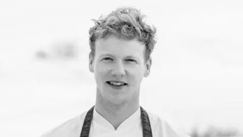 Handout Tommy is smiling. He has blonde curly hair and is wearing a white top with an apron over it. 