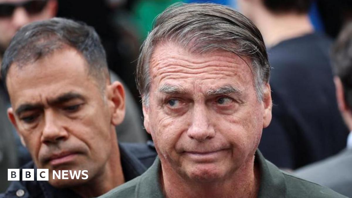 Brazil's former president Jair Bolsonaro is captured mid-wave, his fingers just visible, looking into the distance. His hair is combed over to the right and he is wearing a green khaki shirt.