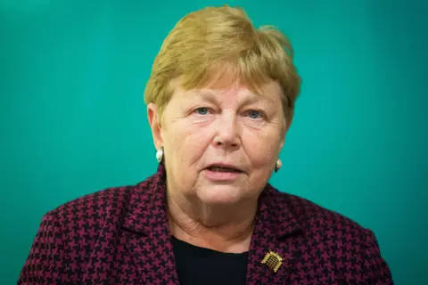 PA Media Baroness Randerson is wearing a dark purple and black houndstooth-patterned blazer over a black top. A small decorative brooch shaped like a building or structure is pinned to the left side of the blazer. She also has pearl earrings. The background is a solid teal colour, creating a clean and simple backdrop.