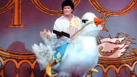 King's Theatre Gerard Kelly performing in panto. He a middle aged man with dark hair, riding a giant goose prop while holding a book in one hand. 