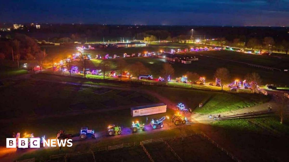 A drone shot of brightly-lit trucks and tractors snaking along a road that zig-zags through green fields, with the lights stretching into the middle distance under a dark-blue evening sky.