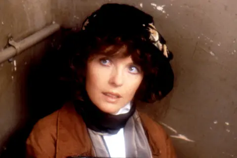 Getty Images Diane Keaton photographed in costume during the filming of the 1980 film Reds. She is seated in a small, enclosed space with beige walls and a visible metal pipe running along the left side. She is wearing a brown coat over a white garment and has a dark hat adorned with a light-coloured patterned scarf or ribbon. The lighting is dim, creating a shadowy and confined atmosphere.
