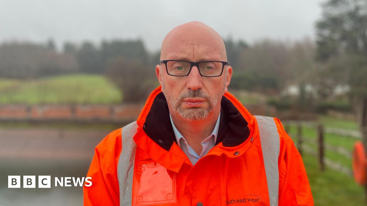 A man in a high-vis orange jacket that says South East Water on it. A body of water and some trees can be seen in the blurry background. He has a stern expression.